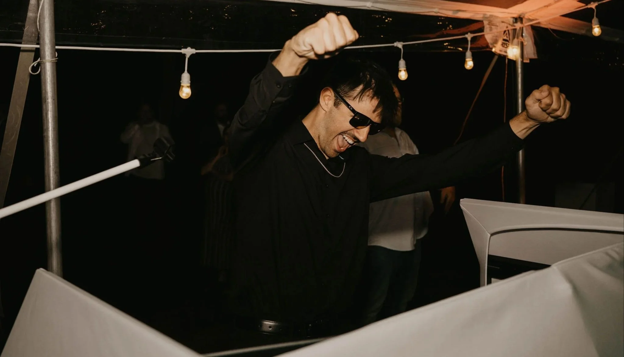 A DJ wearing sunglasses and a black shirt, smiling and with fists raised, playing a white grand piano at night under hanging light bulbs.