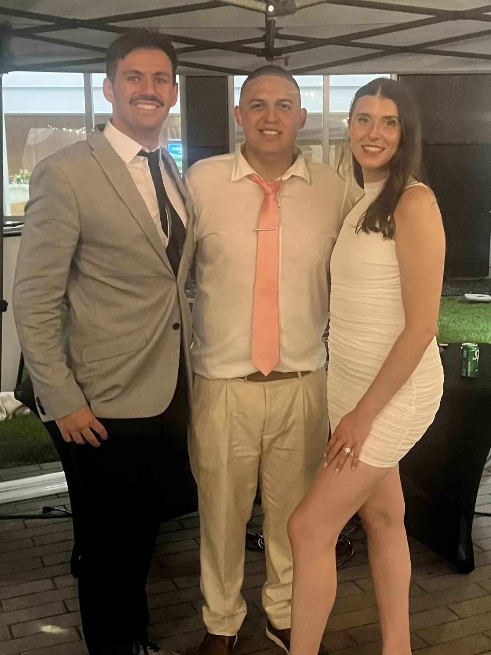 Three people smiling, two men and one woman, standing together at a social event under a canopy, dressed in semi-formal attire, with a background of windows and outdoor evening lighting.