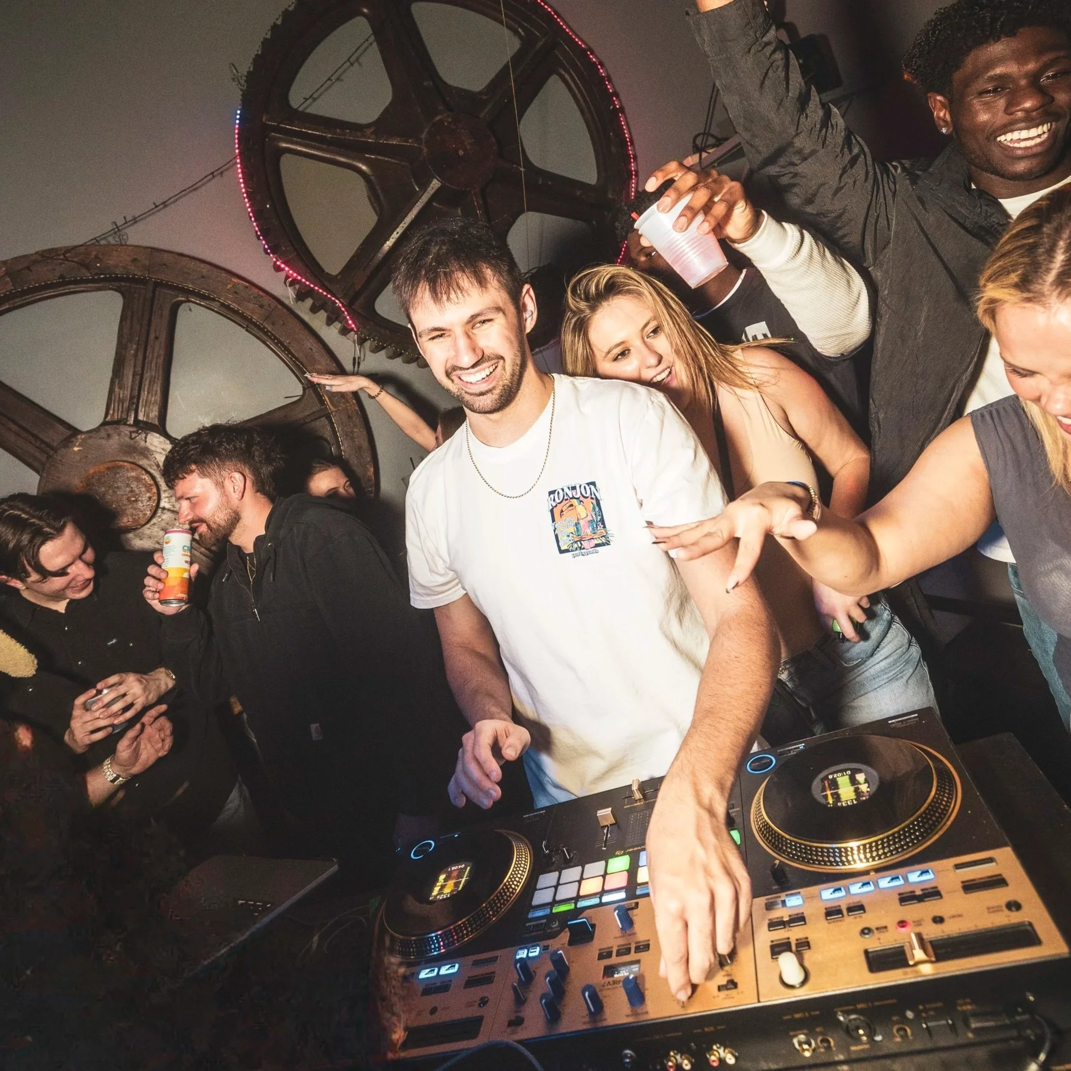 People enjoying a party with Alby playing music, smiling, dancing, and holding drinks in a dimly lit room decorated with large metallic wheels on the wall.