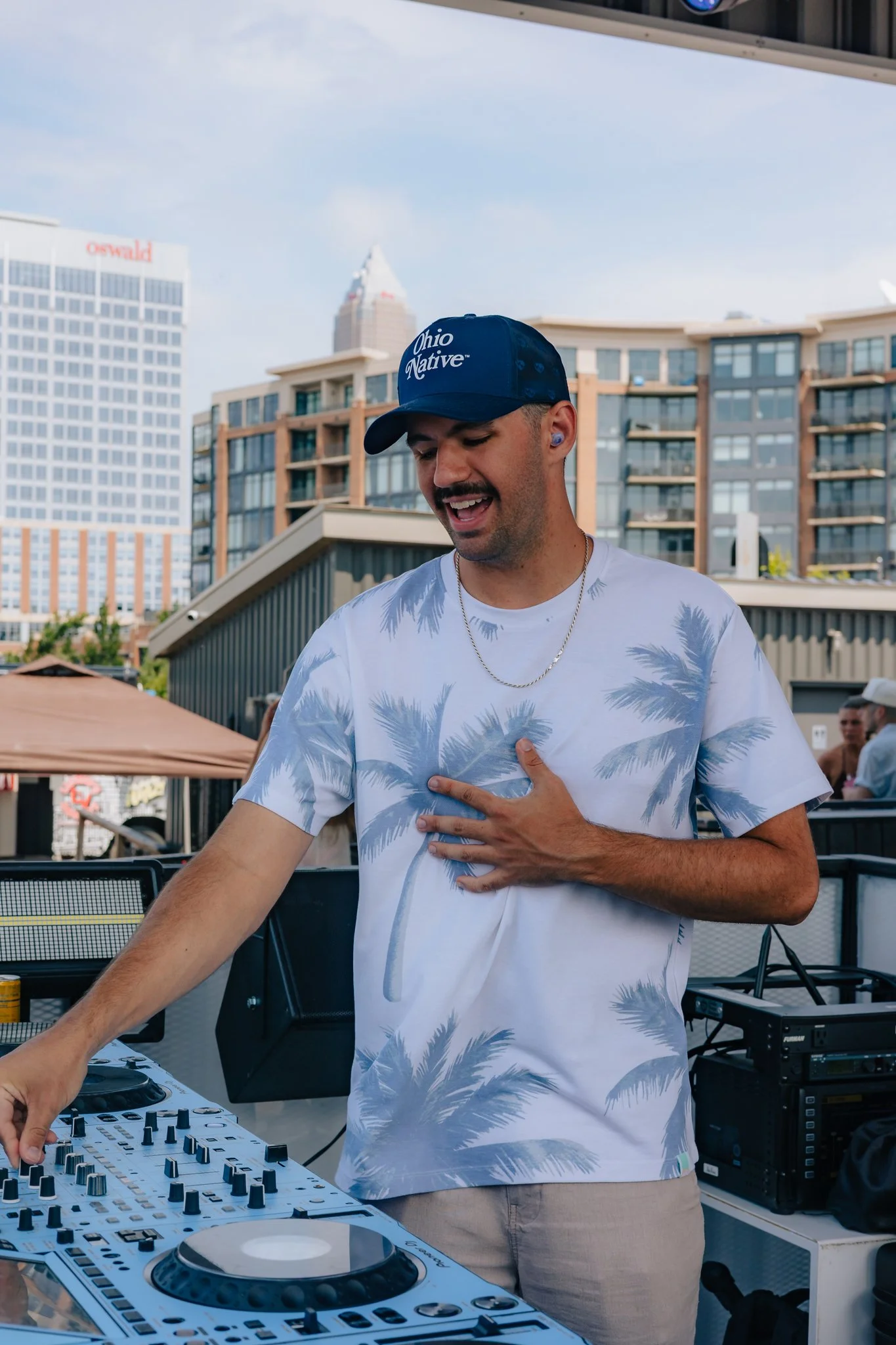 A man wearing a blue cap with the words ''Ohio Native'' and a white t-shirt with a palm tree pattern, DJing at an outdoor event with DJ equipment, in a city with modern buildings in the background.
