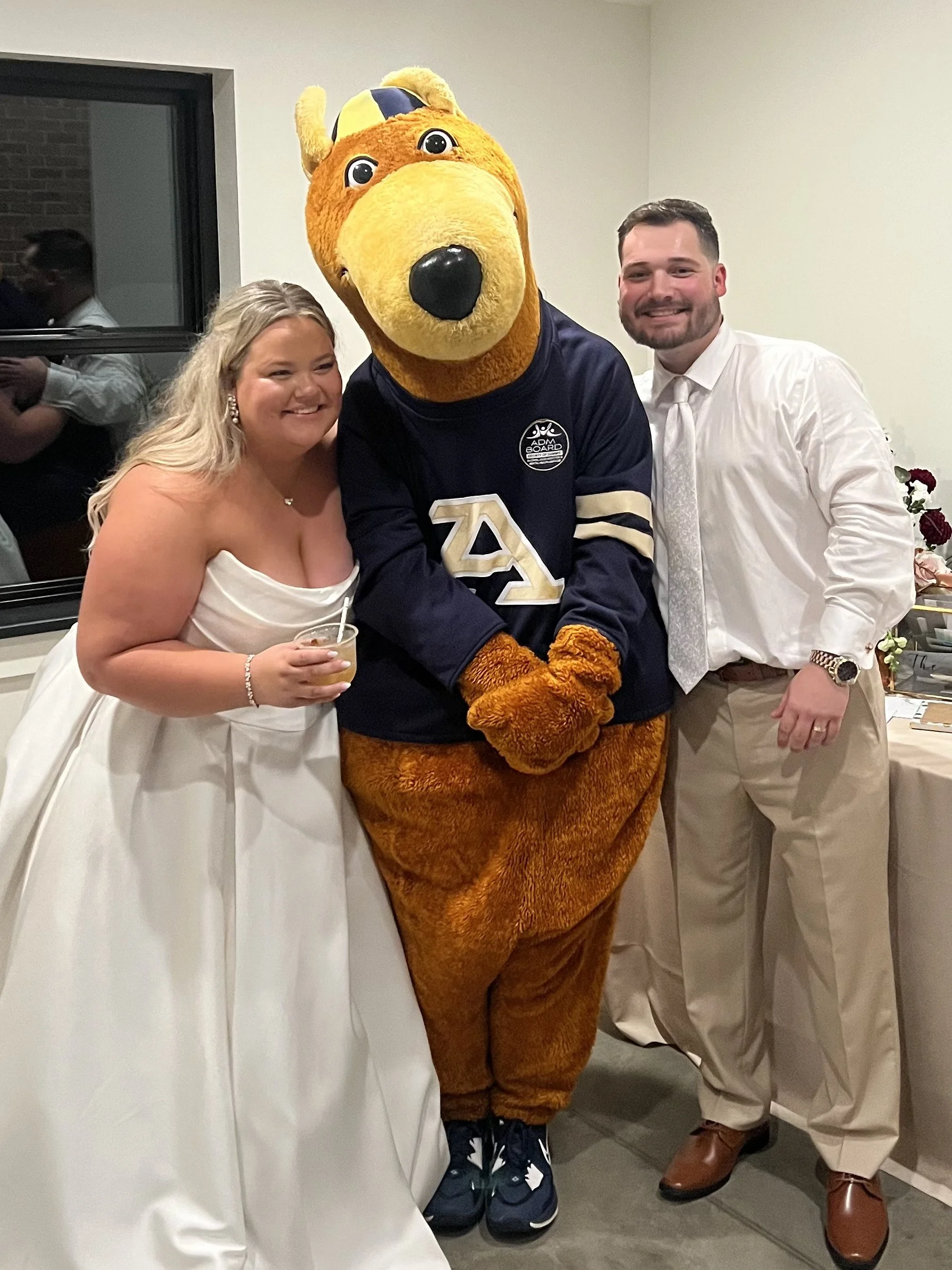 A bride, Zippy, the University of Akron's Mascot, and a groom posing together indoors, smiling for the camera.