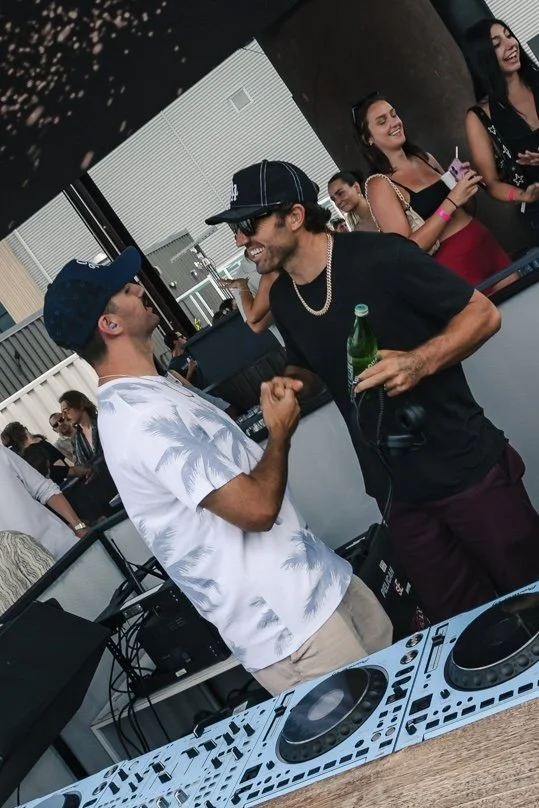 Two men shaking hands at a music event, with two women smiling in the background and a DJ setup in the foreground.