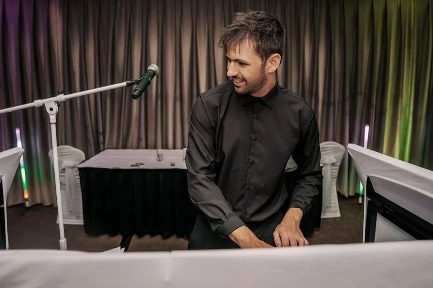 A man with dark hair and a beard, wearing a black shirt, playing keyboard at an event with a curtain backdrop and microphone stand nearby.