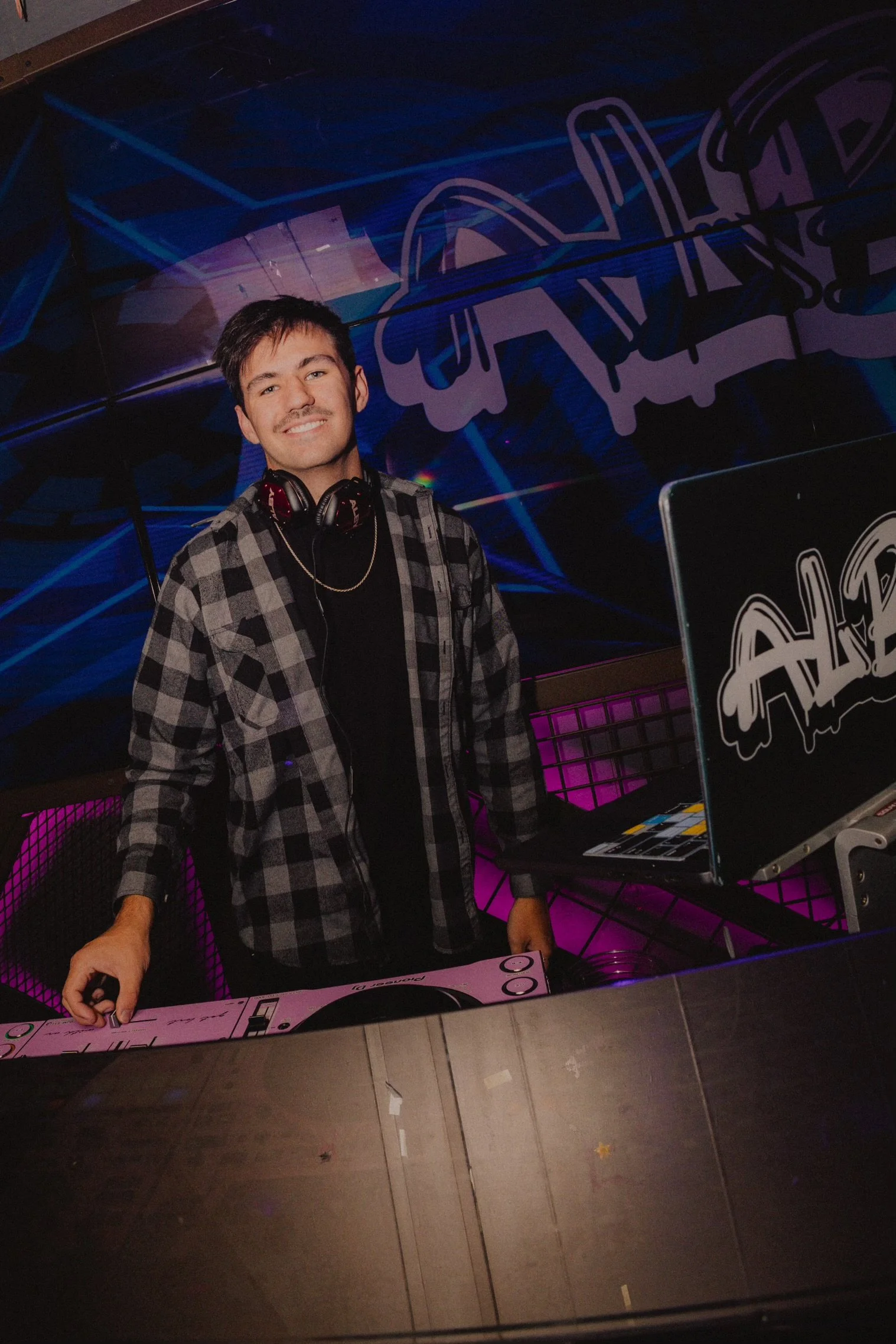 A male DJ wearing headphones and a black and gray plaid shirt standing behind a DJ booth with equipment, smiling at the camera in a nightclub with neon purple and blue lighting and graffiti-style wall art.
