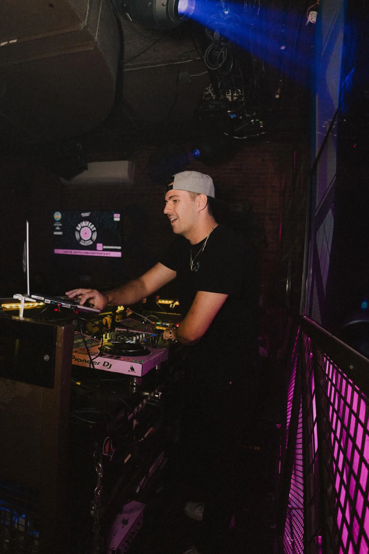 A DJ performing at a nightclub with electronic equipment, wearing a black t-shirt, a baseball cap, and a gold necklace.