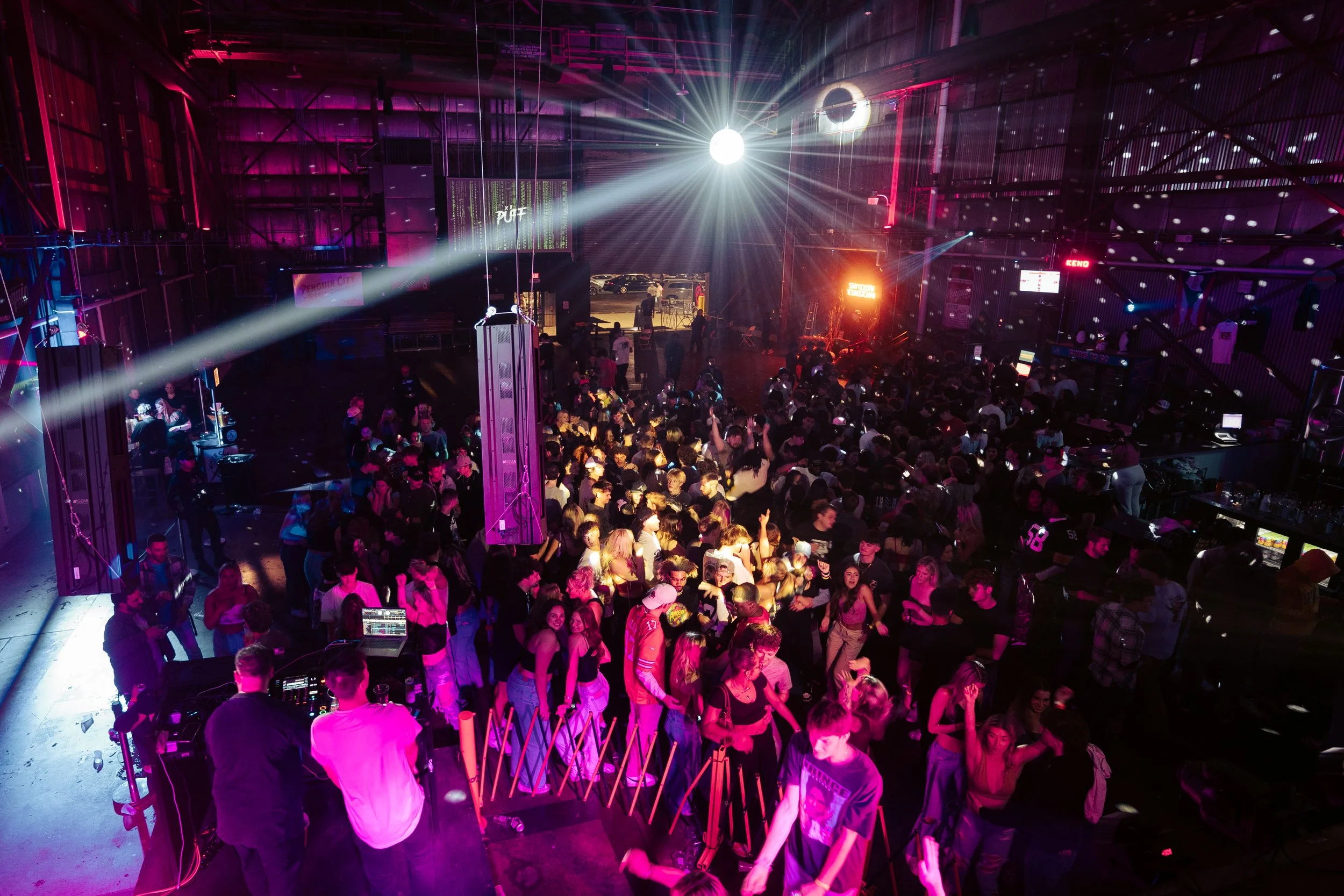 A crowded indoor dance club with colorful lighting, a DJ booth in the foreground, and a disco ball shining bright on the ceiling.