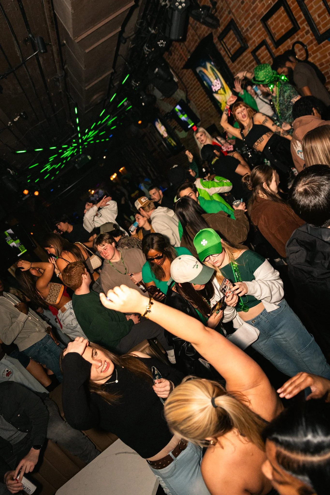 People dancing and socializing at a nightclub with green laser lights, colorful decorations, and a brick wall in the background.