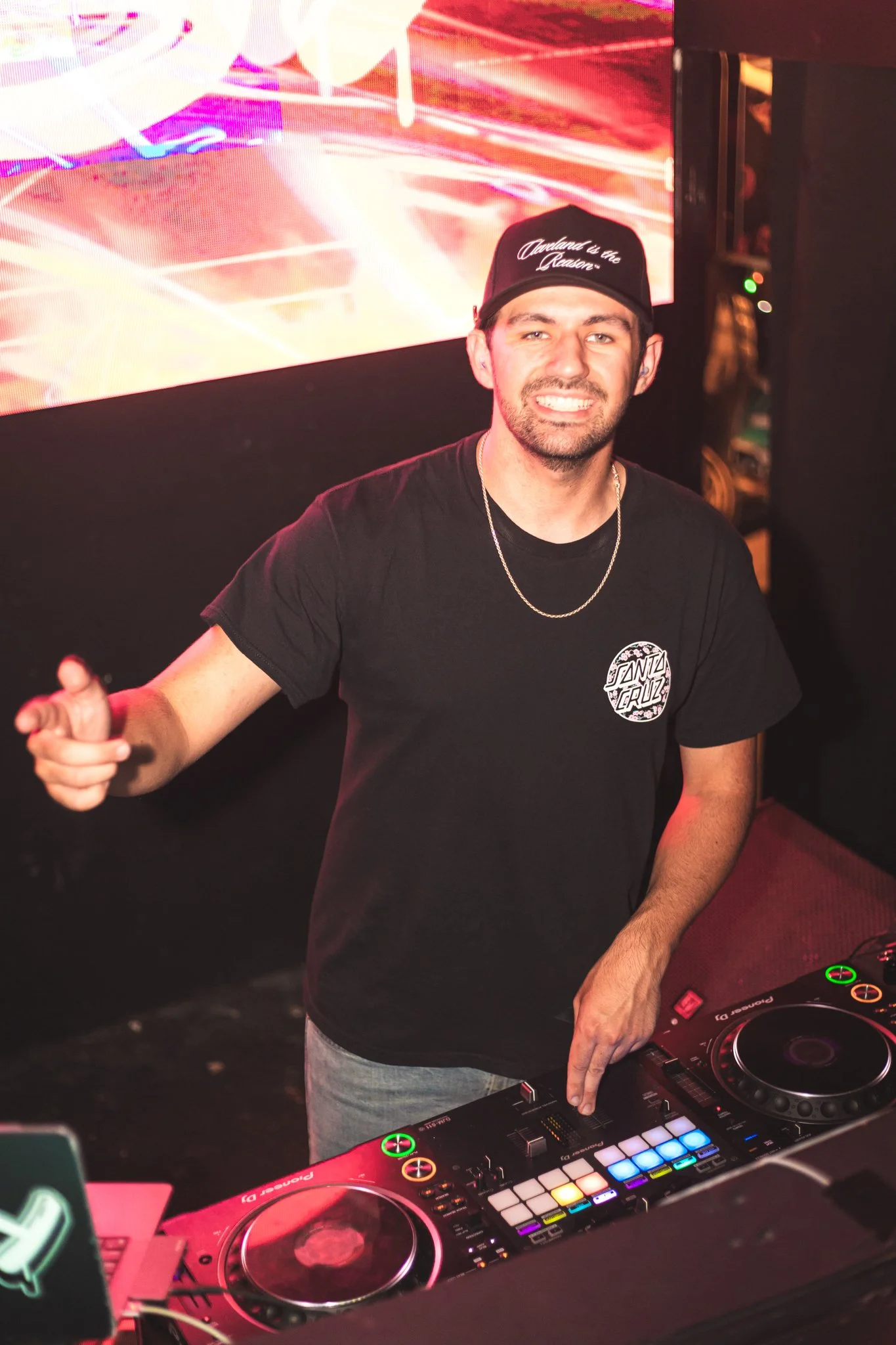 Alby smiling in front of DJ equipment at a nightclub or party, wearing a black T-shirt with a logo, a chain necklace, and a black cap that says "Cleveland is the Reason".