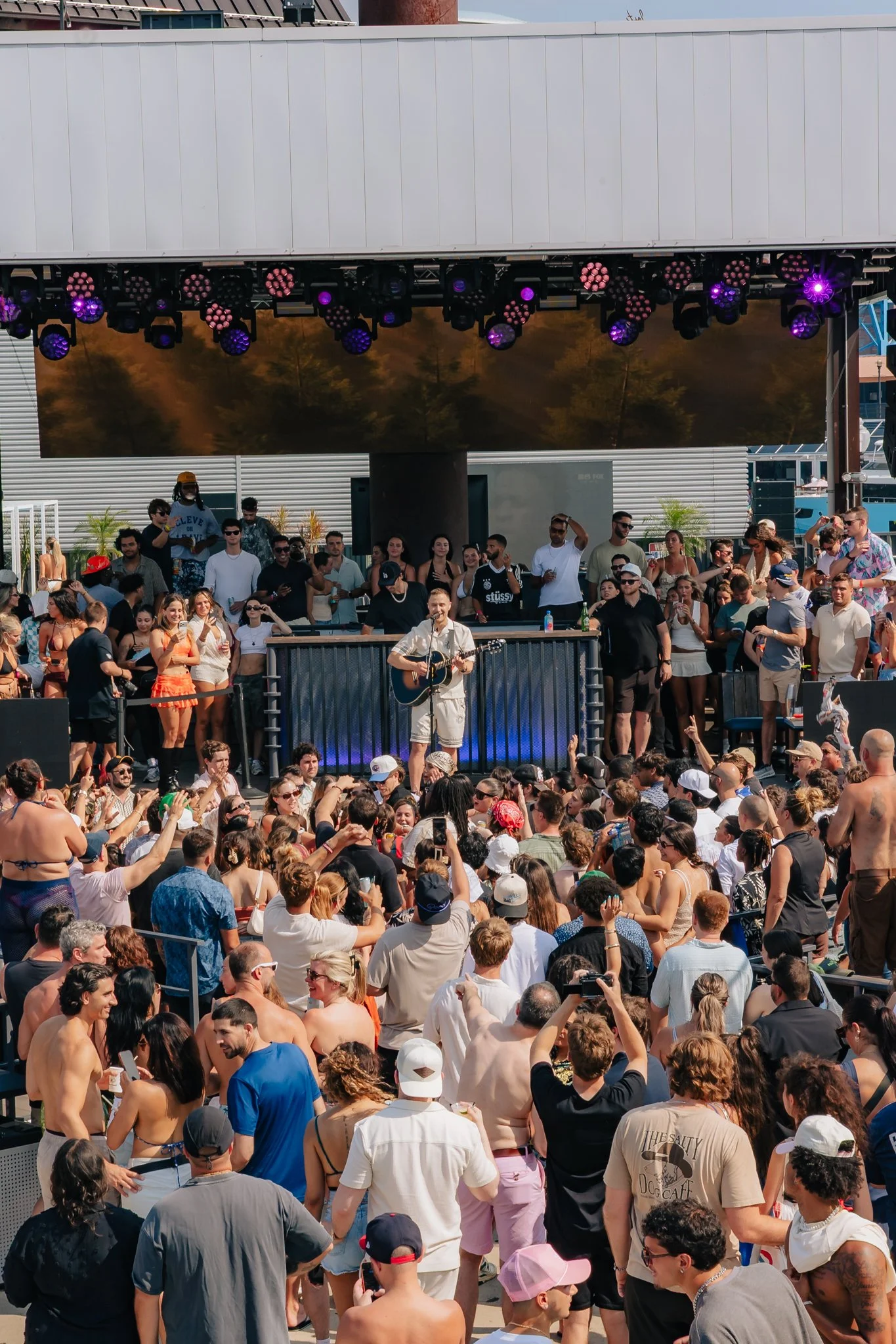 A large crowd of people gathered outdoors at a concert during the daytime. A musician is performing on stage with a guitar. Stage lighting is visible above the stage, illuminating the scene.