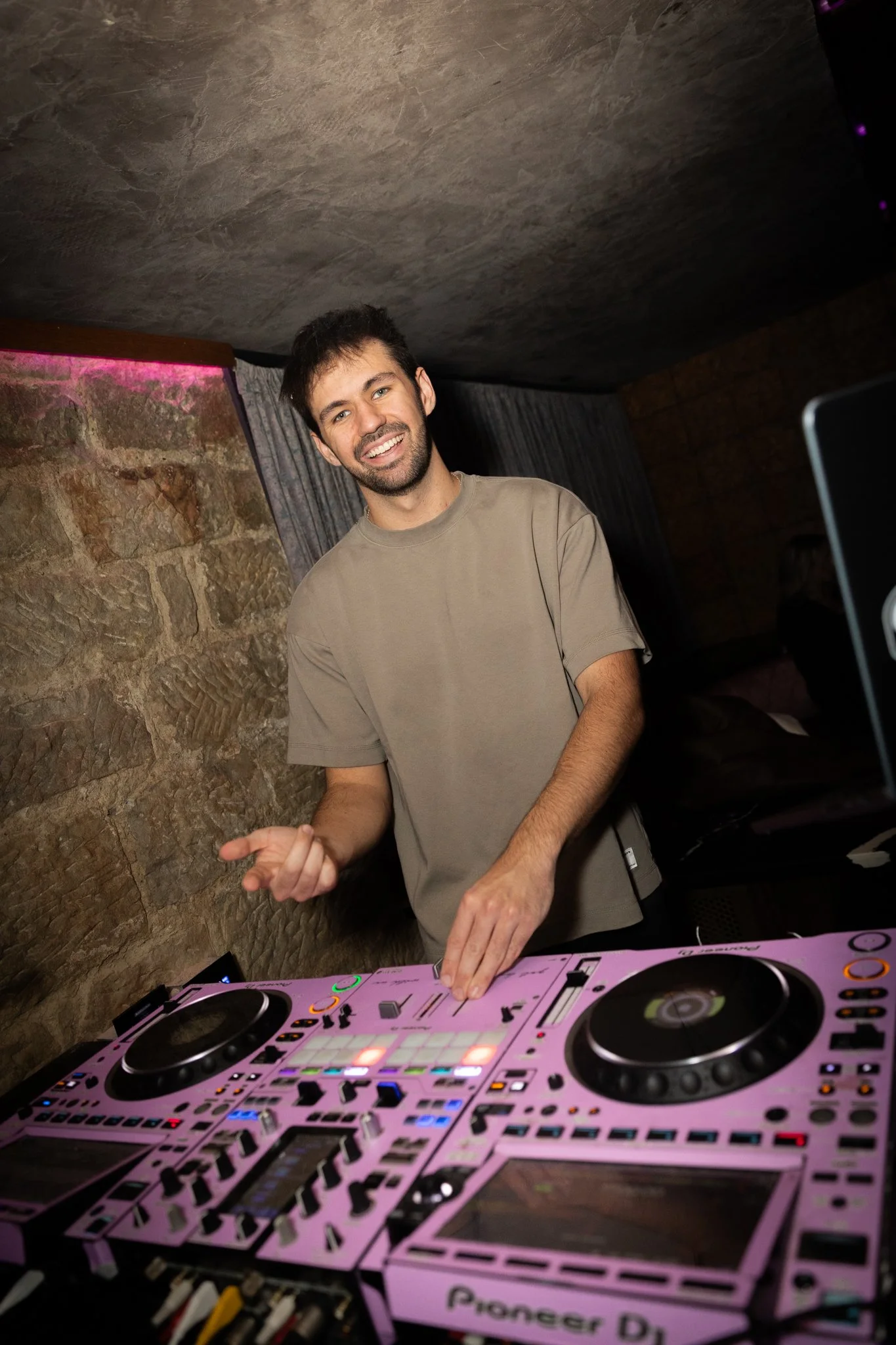 A young man with dark hair and beard smiling while DJing at a party, standing behind a pink Pioneer DJ controller, in a dimly lit room with stone walls and curtains.