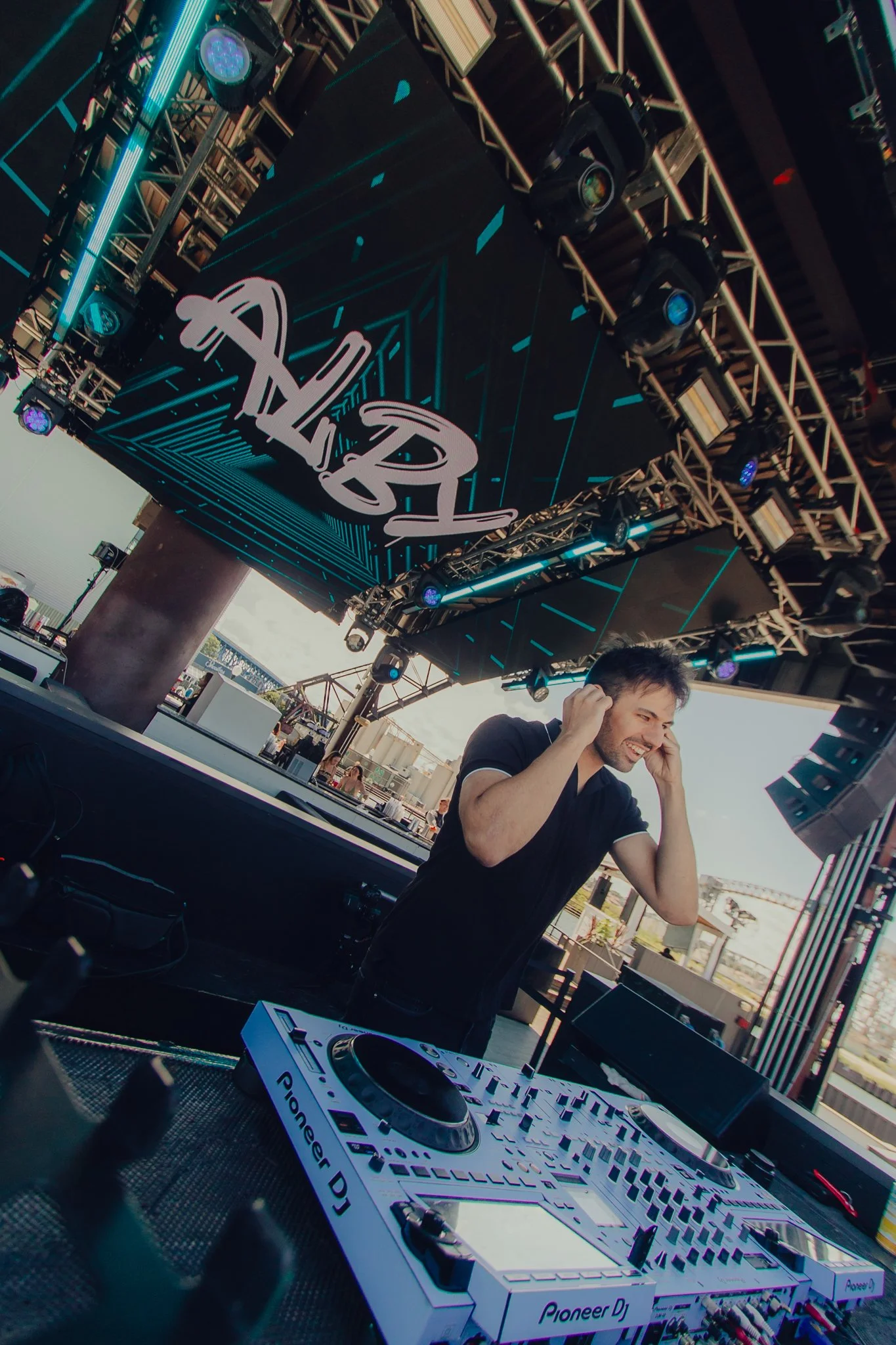 A DJ is performing at an outdoor event, smiling and adjusting his headphones. He is standing behind a Pioneer DJ controller with a large LED screen overhead displaying the word 'ALIEN' in stylized text. The stage is equipped with various lighting and