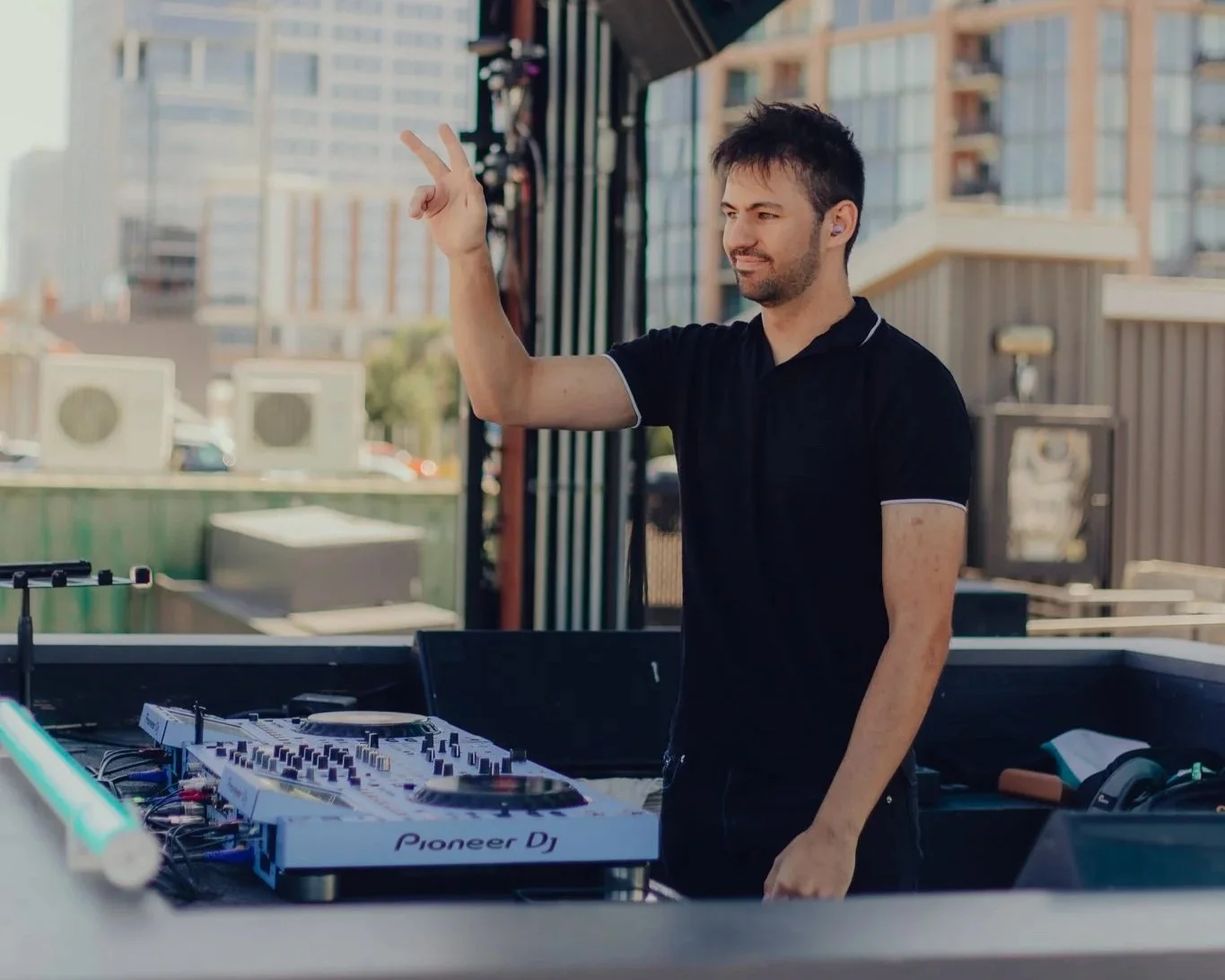 Alby DJing outdoors at FWD Day + Night Club with city buildings in the background, wearing a black shirt and making a peace sign with his right hand.