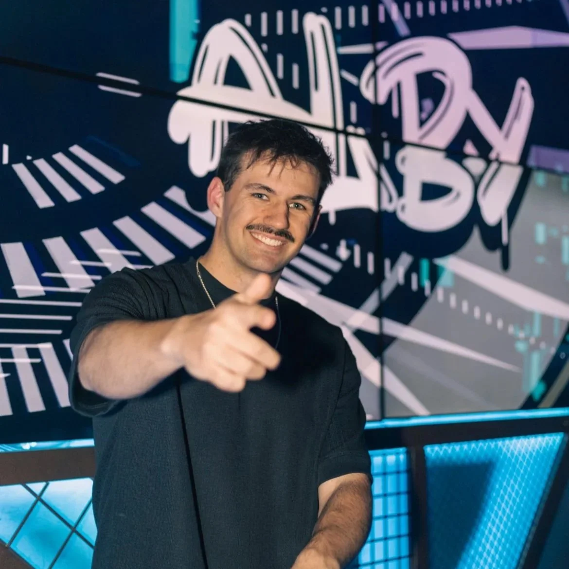 Alby smiling and pointing at the camera in front of a colorful LED display with his logo in an abstract graffiti-style writing.