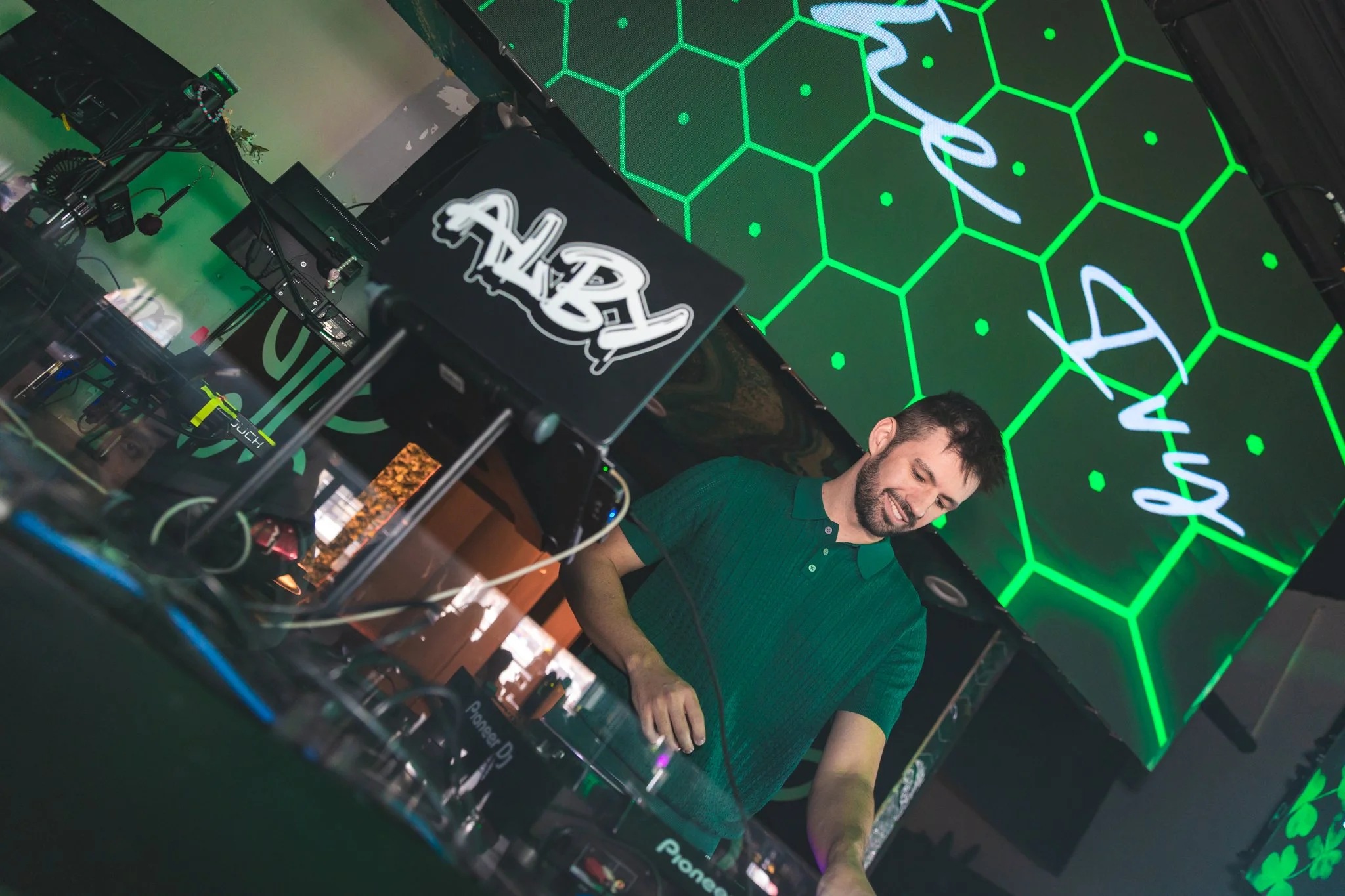 A DJ in a green polo shirt is performing at a club, surrounded by DJ equipment and a large screen displaying green honeycomb pattern with the word 'Arom'.