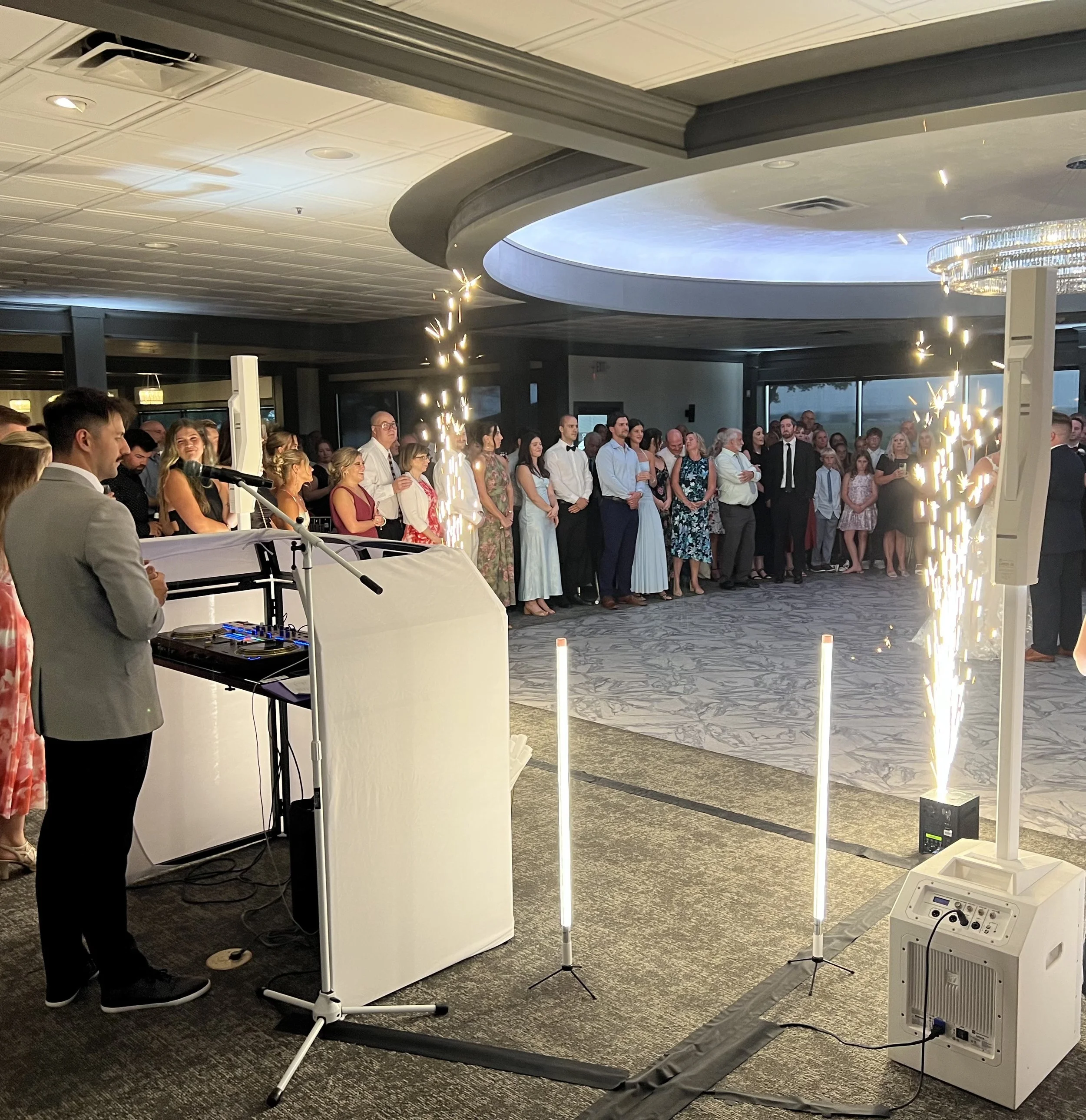 People attending an indoor event with sparklers and a DJ setup, in a decorated hall with a modern ceiling and chandelier.