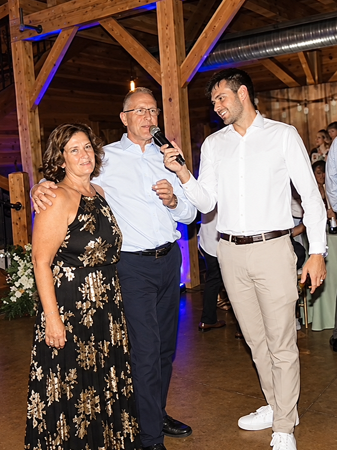 A woman in a black floral dress, a man in a white shirt with glasses, and a young man in a white shirt and beige pants at a wedding reception or celebration inside a wooden venue, with the young man holding a microphone.