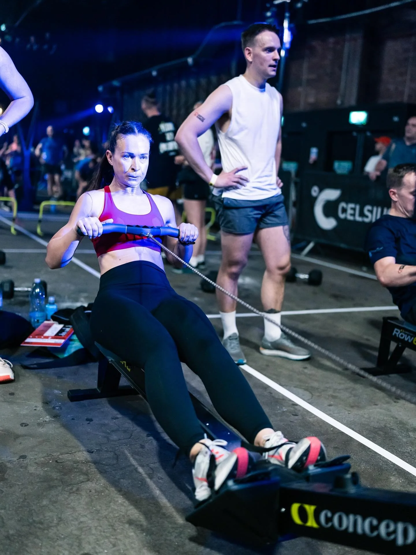A woman using a rowing machine at a fitness event, with a man standing behind her and other people in the background.
