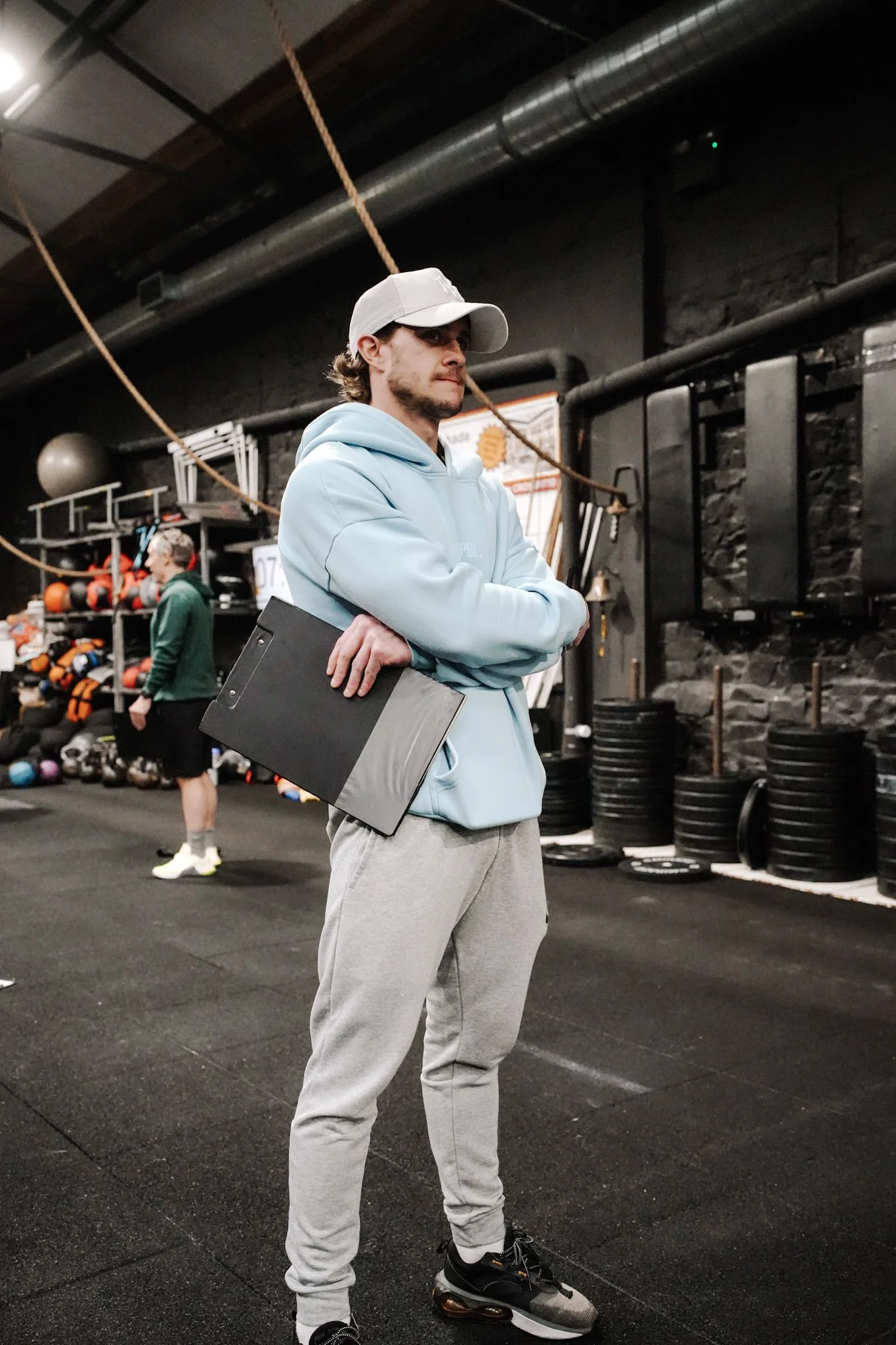 A man in a light blue hoodie and gray sweatpants standing in a gym, holding a clipboard, with gym equipment and another person in the background.