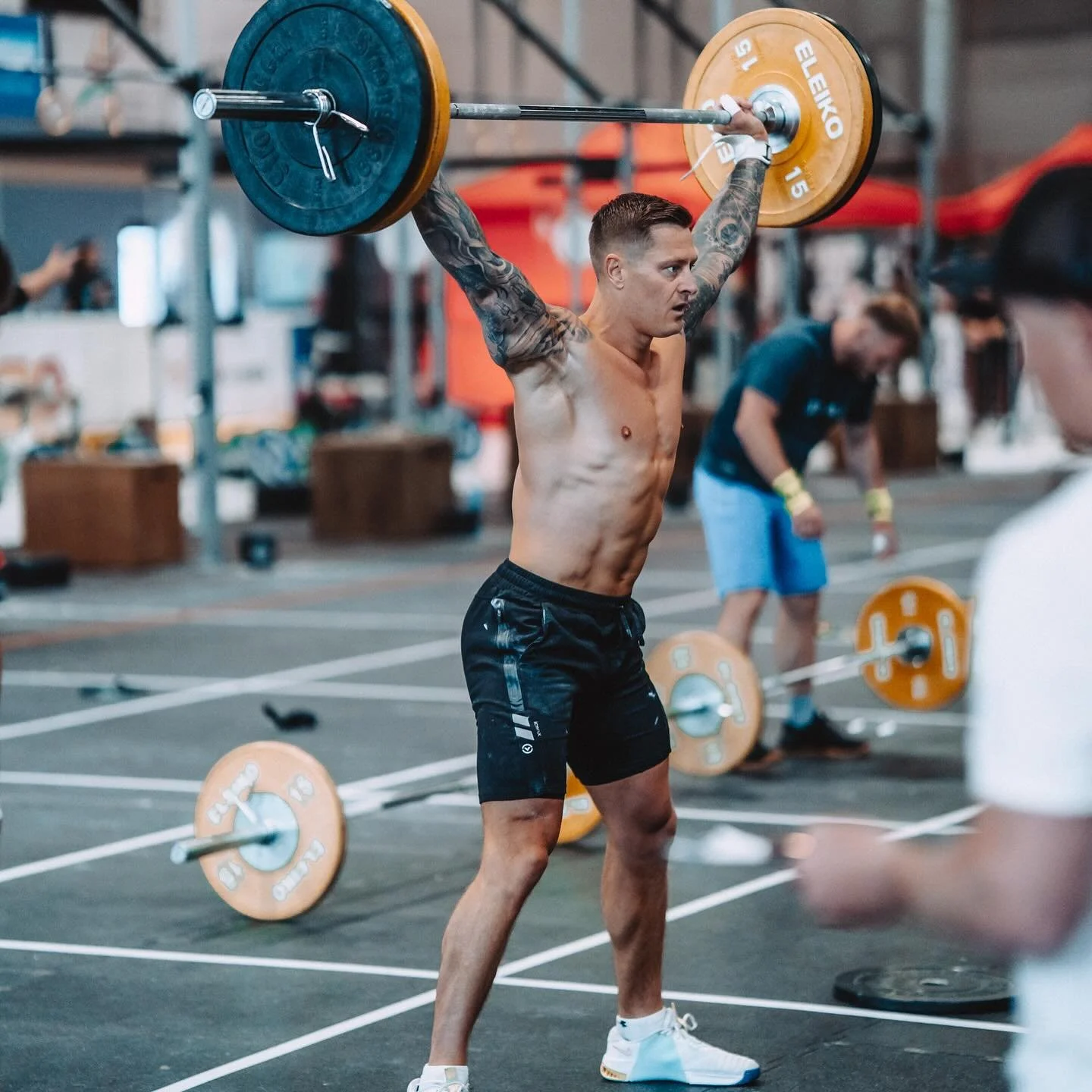 A man lifting a barbell with orange weights overhead during a workout at a gym.