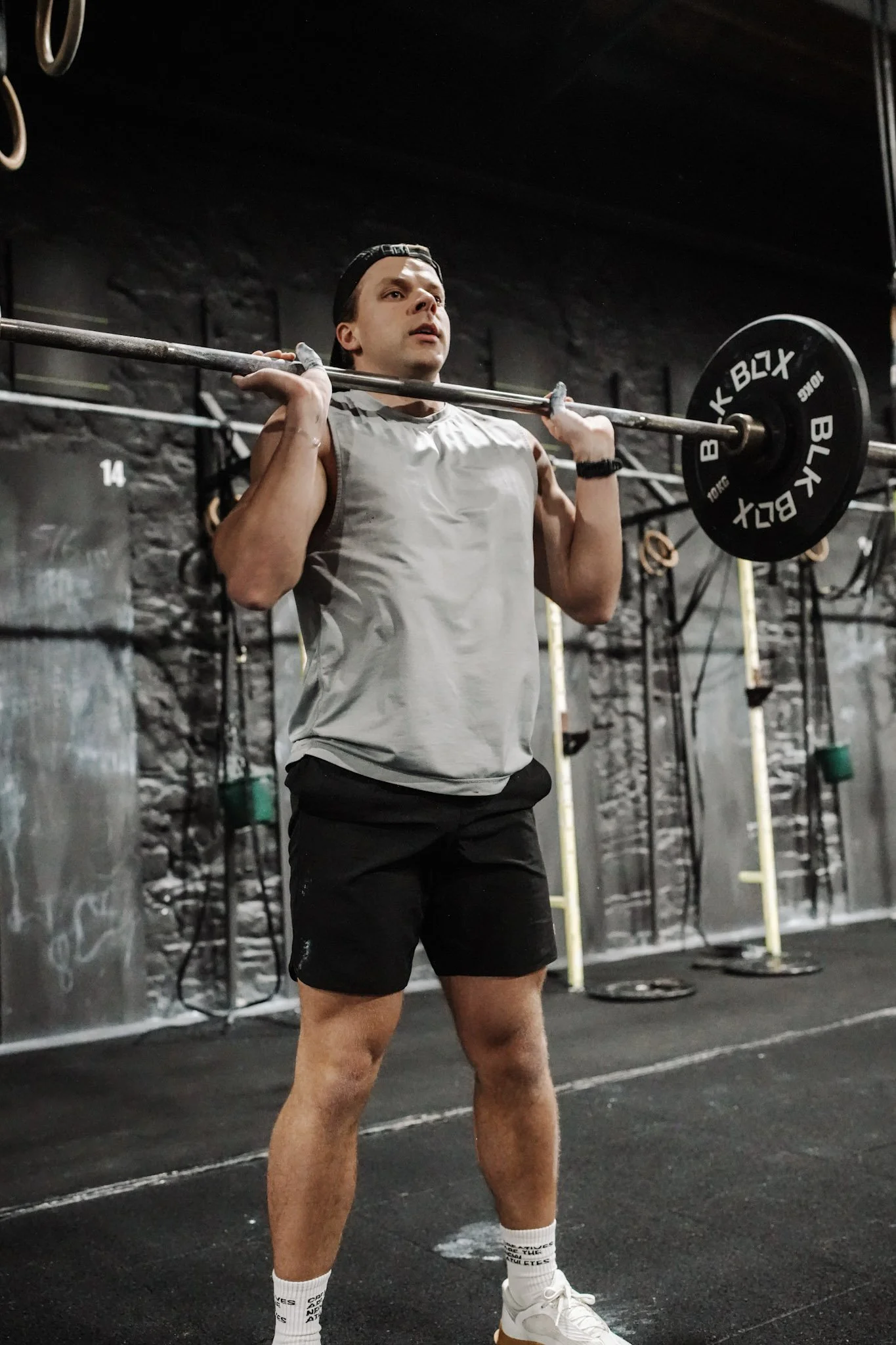 A man lifting a barbell with weight plates in a gym. He is wearing a sleeveless shirt, black shorts, white sneakers, and a black cap, standing on a black rubber gym floor.