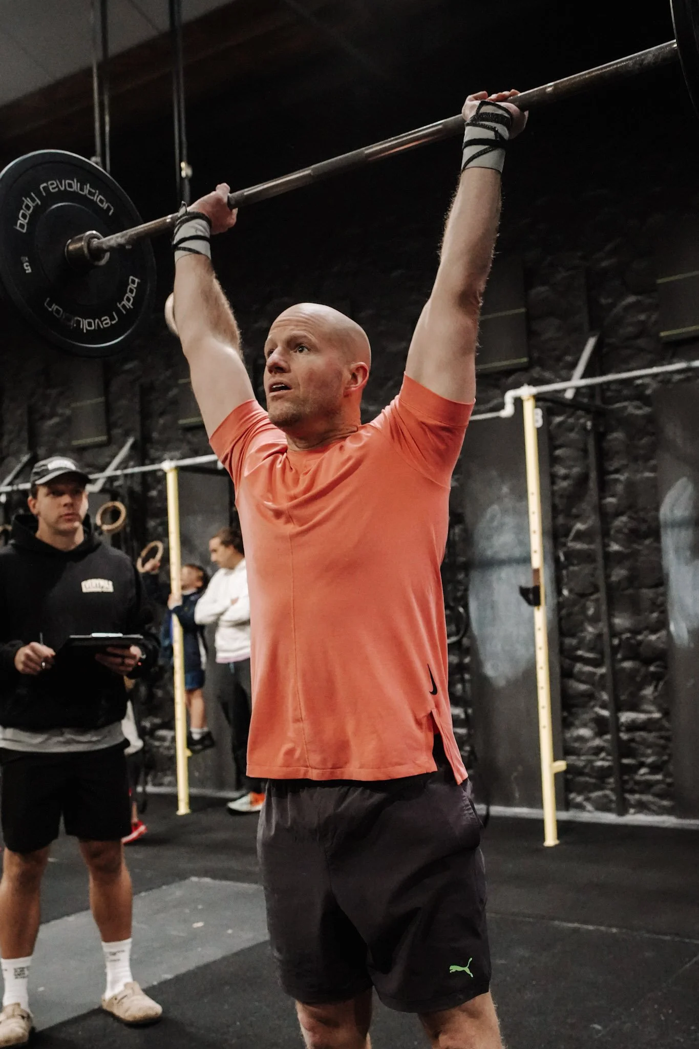 A man wearing an orange shirt and black shorts lifts a barbell overhead during a workout at a gym, with a trainer observing in the background.