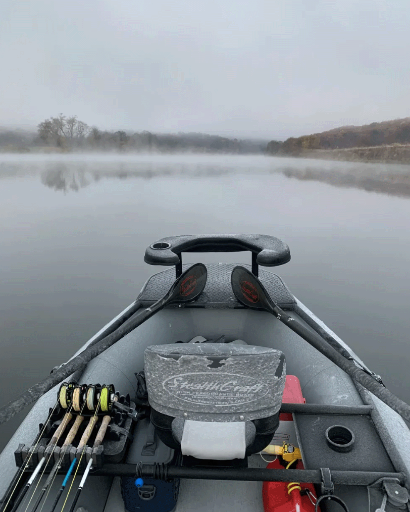 View from a boat on a calm lake with fog, showing the boat's bow with fishing rods and equipment.
