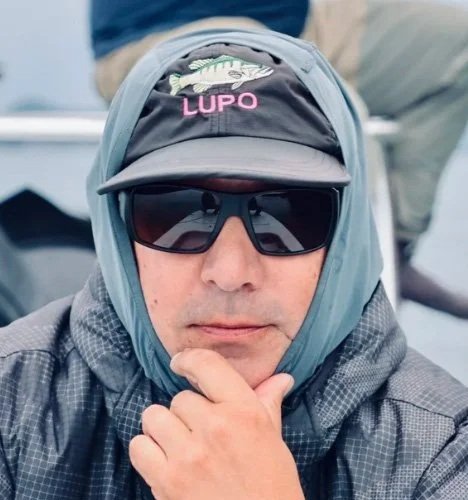 A man wearing a gray hooded jacket, a black cap with 'LUPO' embroidered on it, and dark sunglasses, posing outdoors with his hand on his chin.