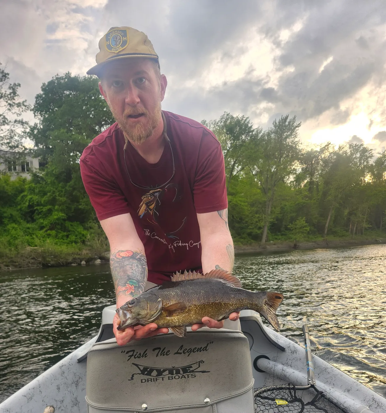 A man with tattoos wearing a beige hat and maroon t-shirt holding a fish on a boat in a river with trees and cloudy sky in the background.