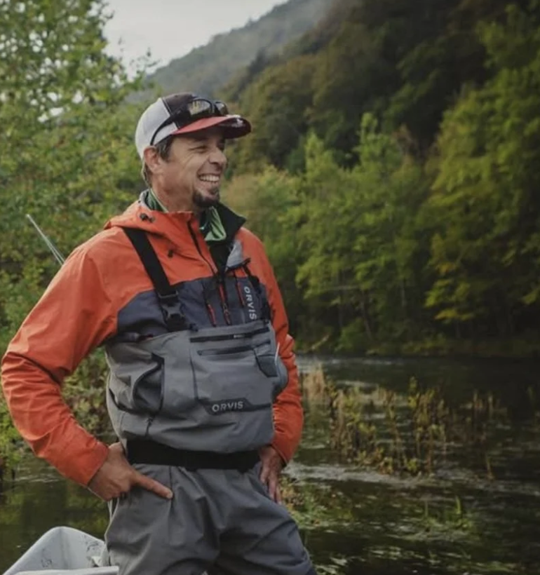 A man wearing outdoor gear, including a red and gray jacket and cap, stands by a river with green trees and hills in the background.