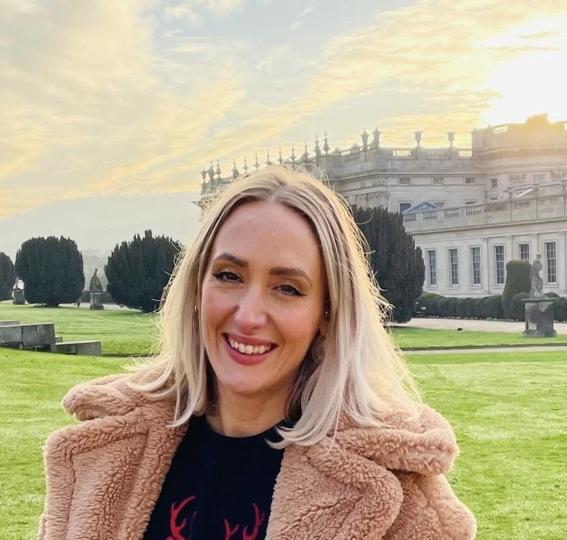A smiling woman with blonde hair standing outdoors in front of a grand historic building with a landscaped garden and cloudy sky.