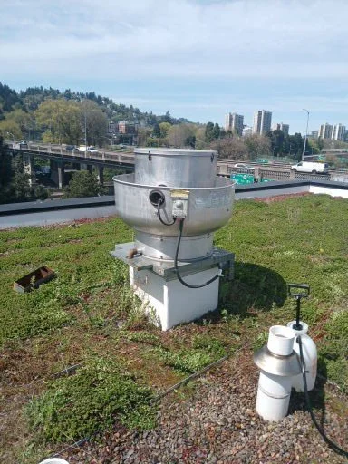 Industrial rooftop ventilation or exhaust fan with control box on a grassy rooftop, cityscape with buildings and highways in the background.
