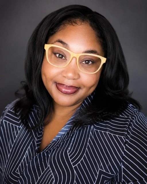 A woman with dark hair wearing yellow glasses and a navy and white striped blouse, smiling against a dark gray background.