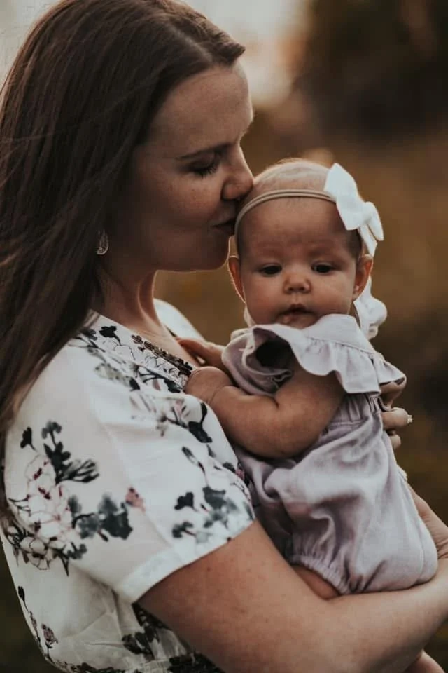 A woman with long brown hair holding a baby girl with a white bow in her hair, outdoor setting during sunset.
