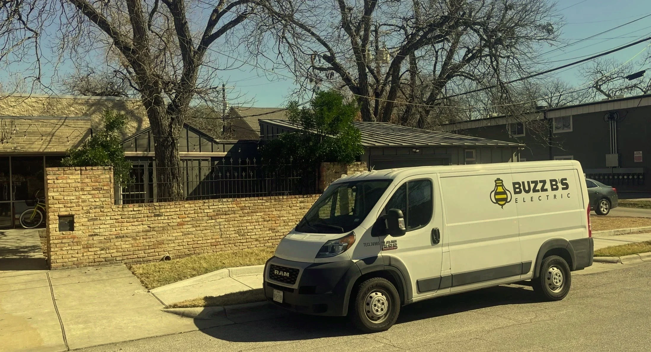 Street view showing part of a residence with a brick wall and metal fence, a tree with no leaves, and a white delivery van labeled Buzz B's Electric parked on the street.