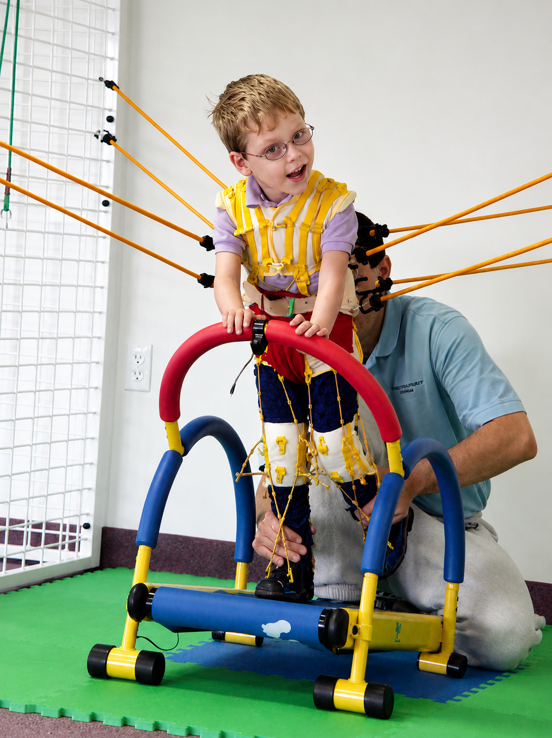 Child with physical therapy harness standing on a grey and blue walk trainer with a green play mat surface, assisted by a therapist in a facility with white walls.