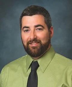 Portrait of a man with dark hair and beard, wearing a green shirt and black tie against a textured blue background.