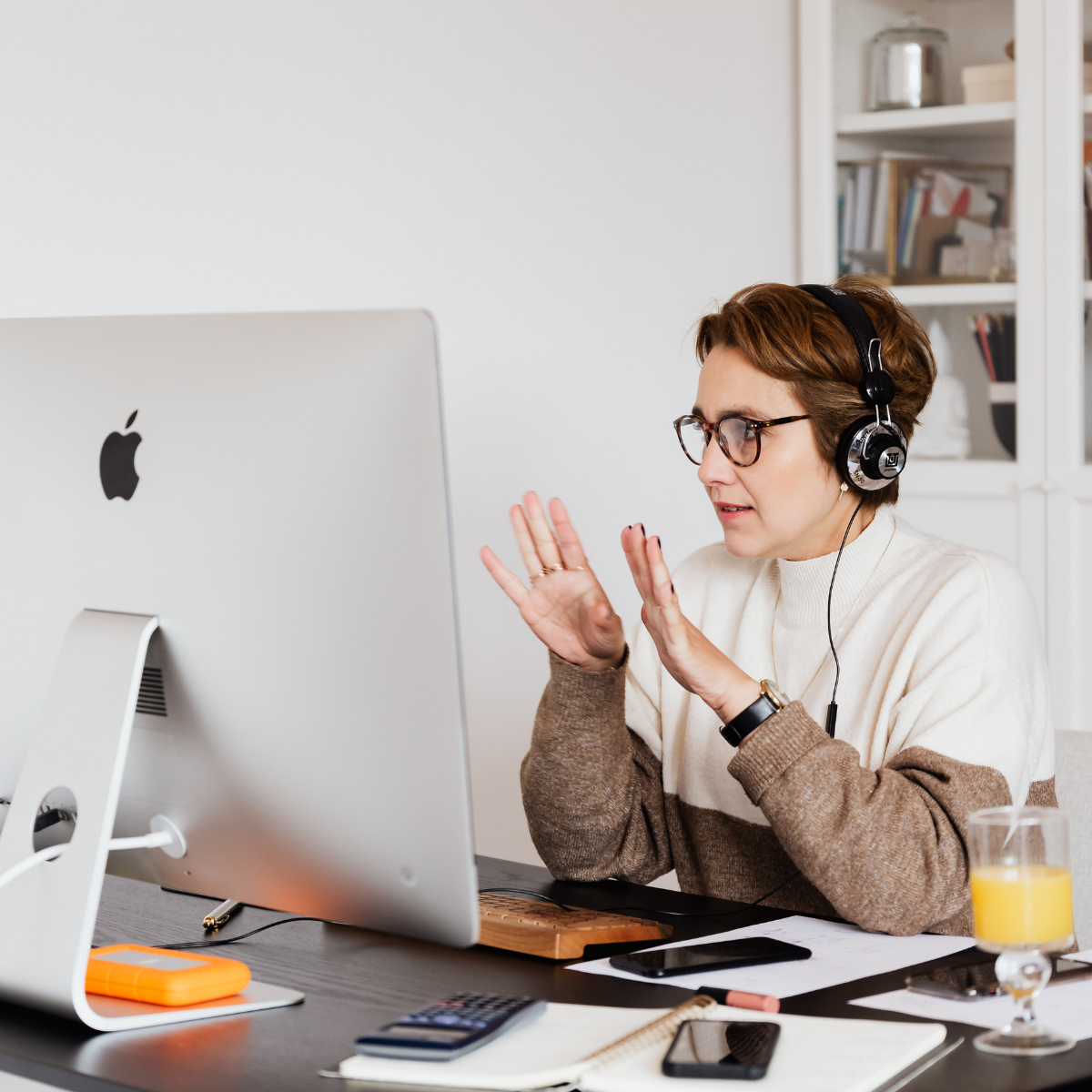 A woman with short brown hair, glasses, and headphones sitting at a desk with an iMac computer, raising her hands as if explaining something. The desk has a glass of orange juice, a smartphone, papers, a calculator, and a pen.