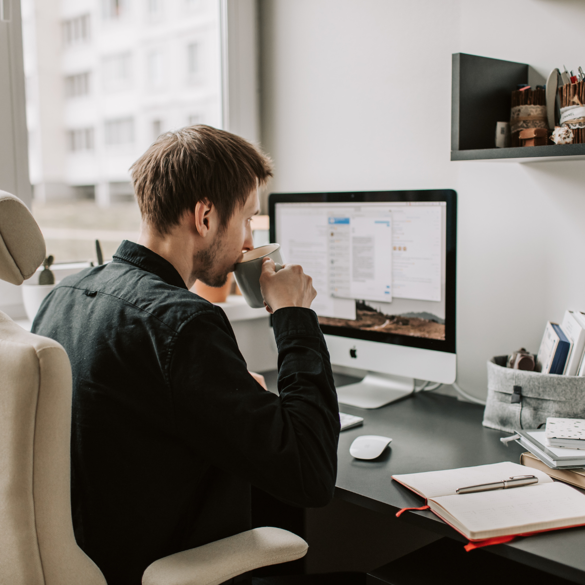 A man sitting at a desk drinking from a mug while working on an iMac computer in a bright room with large windows.