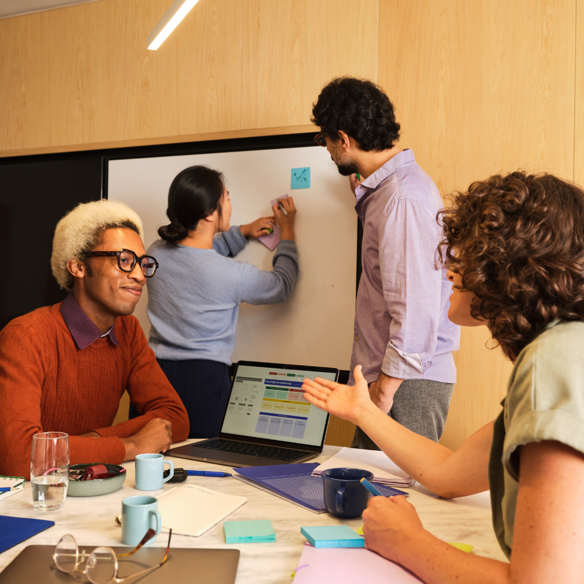 Group of people in a meeting room, some sitting at a table with notebooks, a laptop, and coffee mugs, others standing at a whiteboard with sticky notes, engaged in discussion.