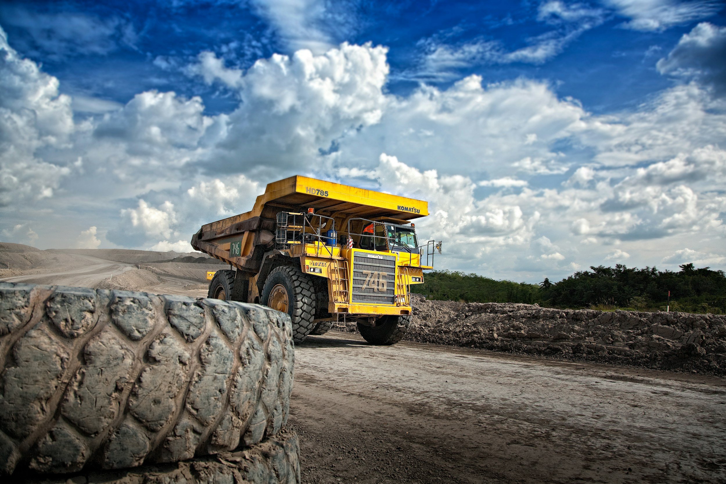 A yellow Komatsu dump truck with black wheels and a yellow bed, driving on a dirt road at a construction or mining site with a cloudy blue sky overhead.