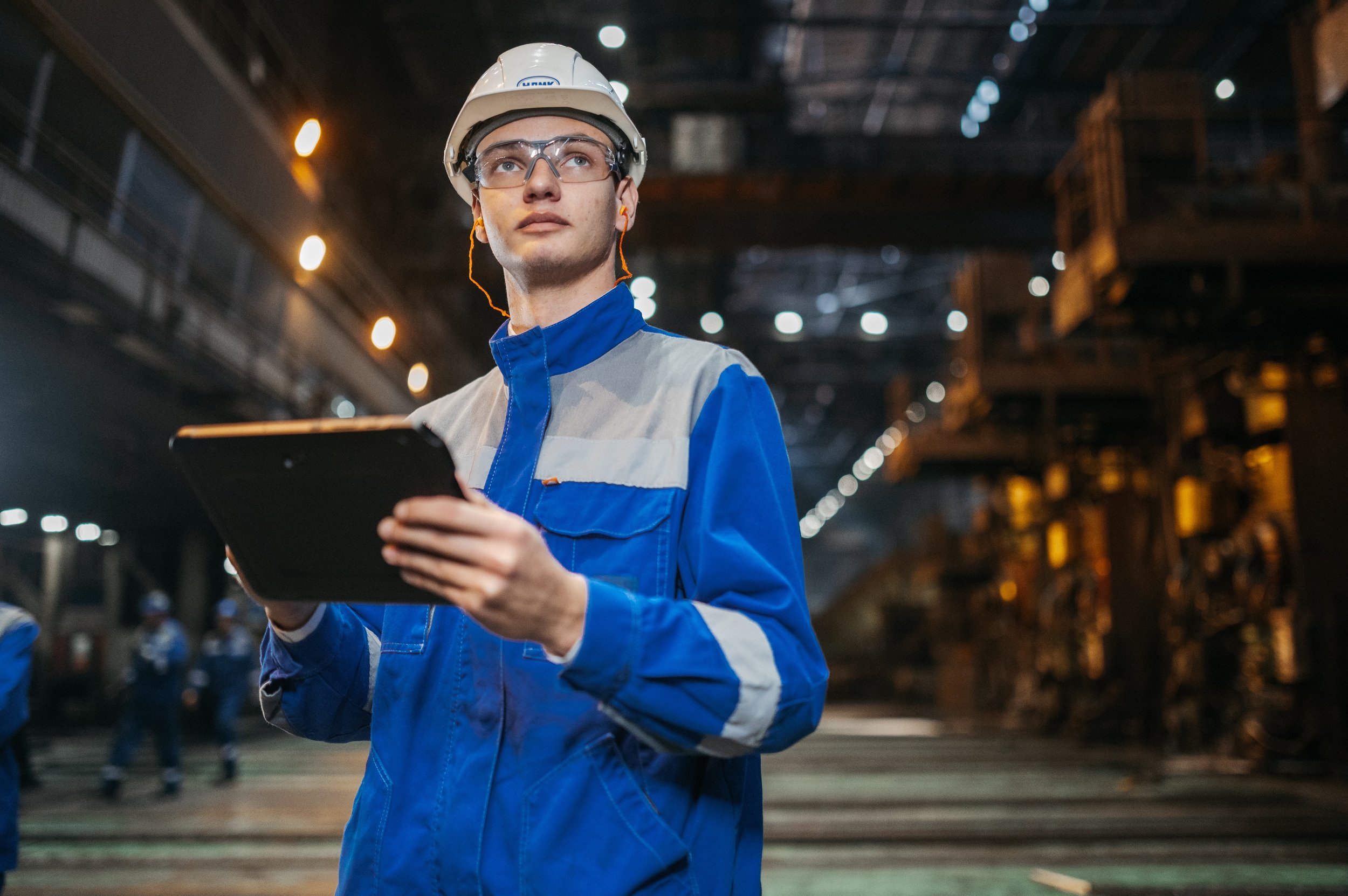 A young male factory worker wearing safety glasses, a hard hat, and a blue uniform holding a tablet, inside an industrial facility with other workers in the background.