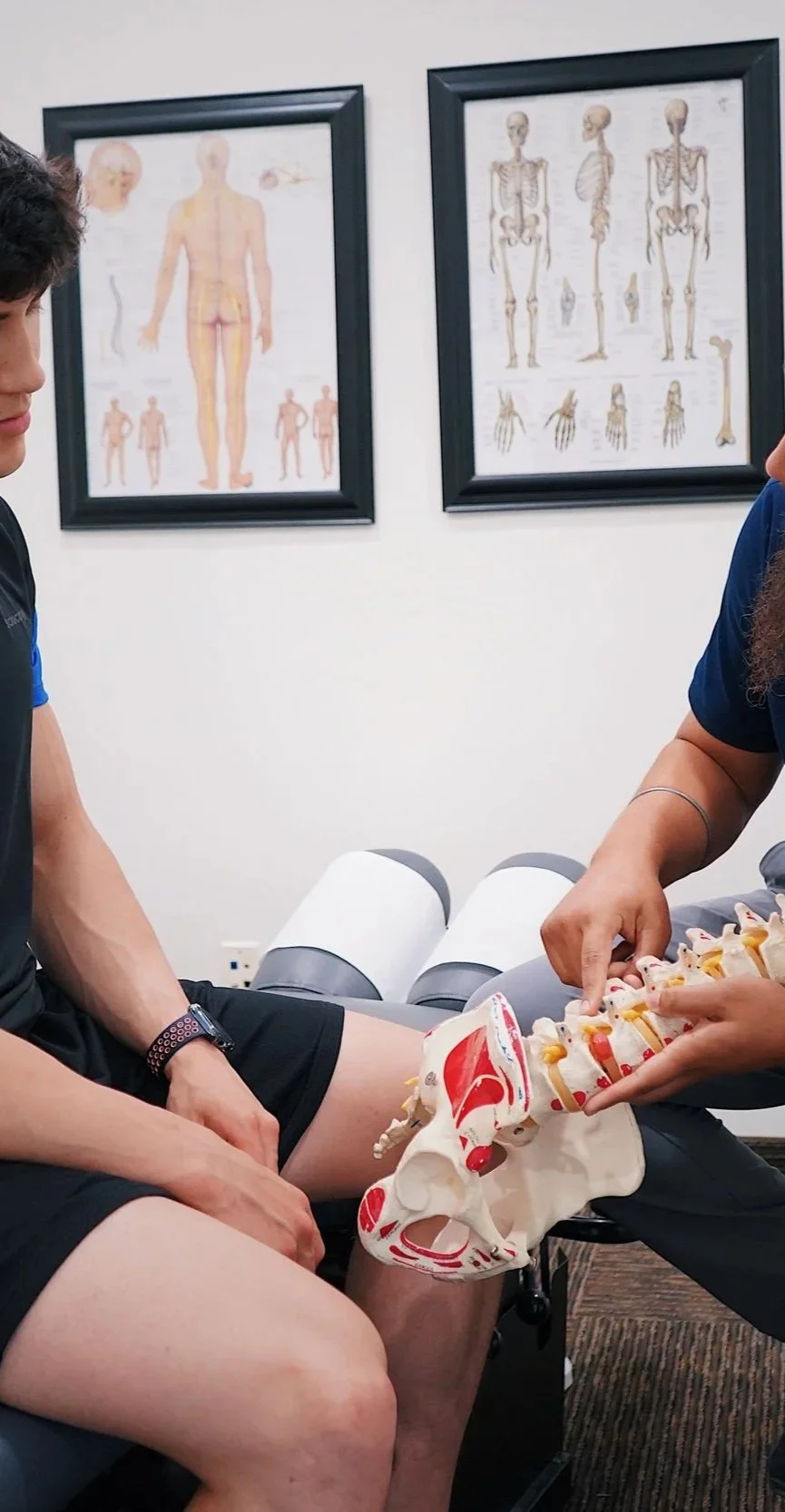 A man sitting in a medical office while a healthcare professional displays a model of the human spine. Art posters of human skeletal and muscular anatomy are on the wall behind.