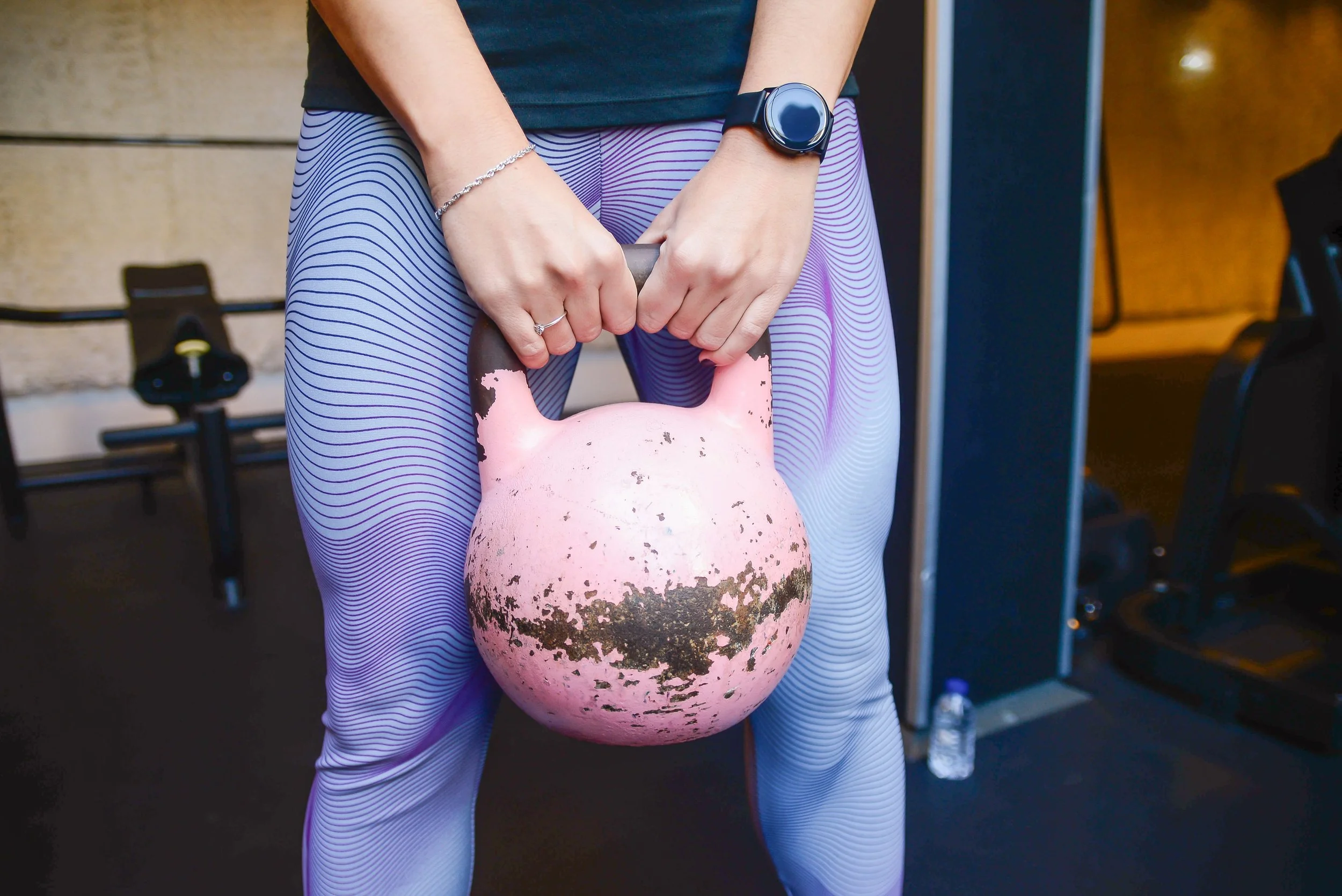 A person holding a large pink and black training kettlebell with both hands. The individual is wearing purple patterned leggings, a black top, a silver bracelet, a ring, and a black smartwatch. The background includes gym equipment and a water bottle.