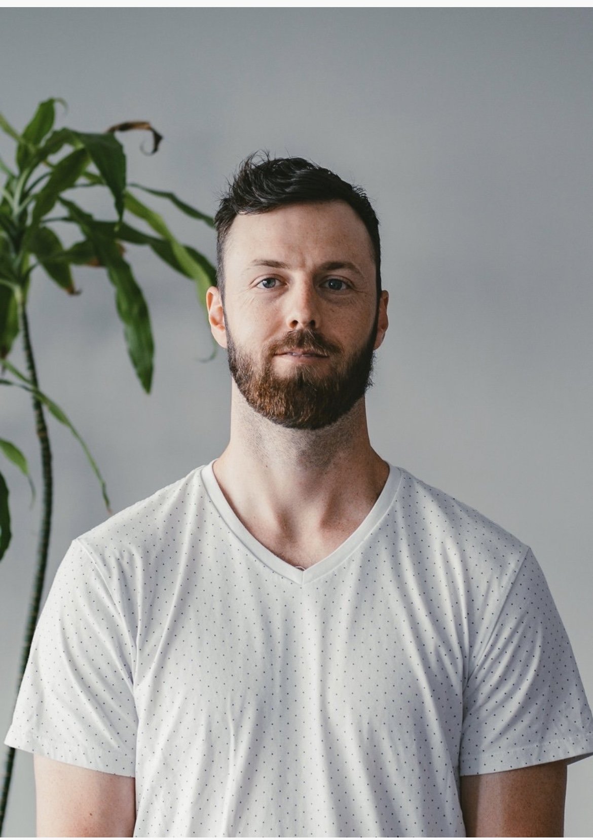 A man with a beard and short dark hair, wearing a white V-neck T-shirt with small dots, standing indoors in front of a gray wall and a green potted plant.