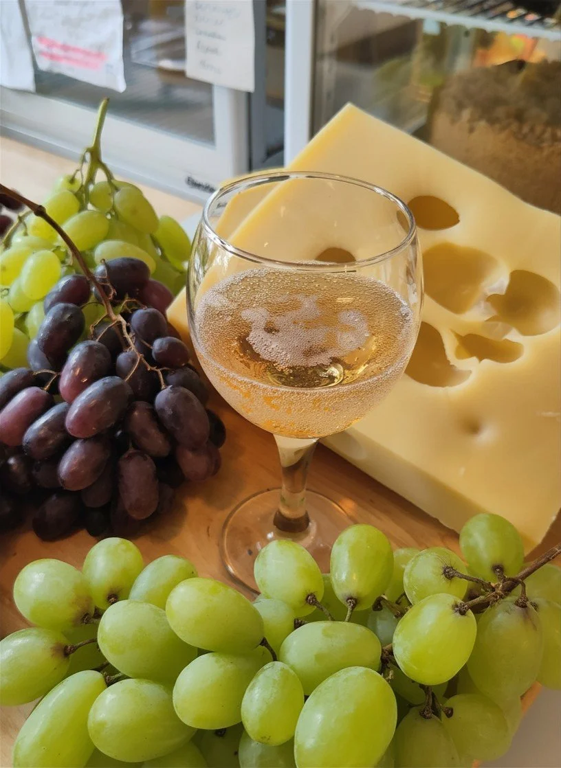 Weißwein im Weinglas, umgeben von grünen und roten Weintrauben, ein Stück Käse mit Löchern.