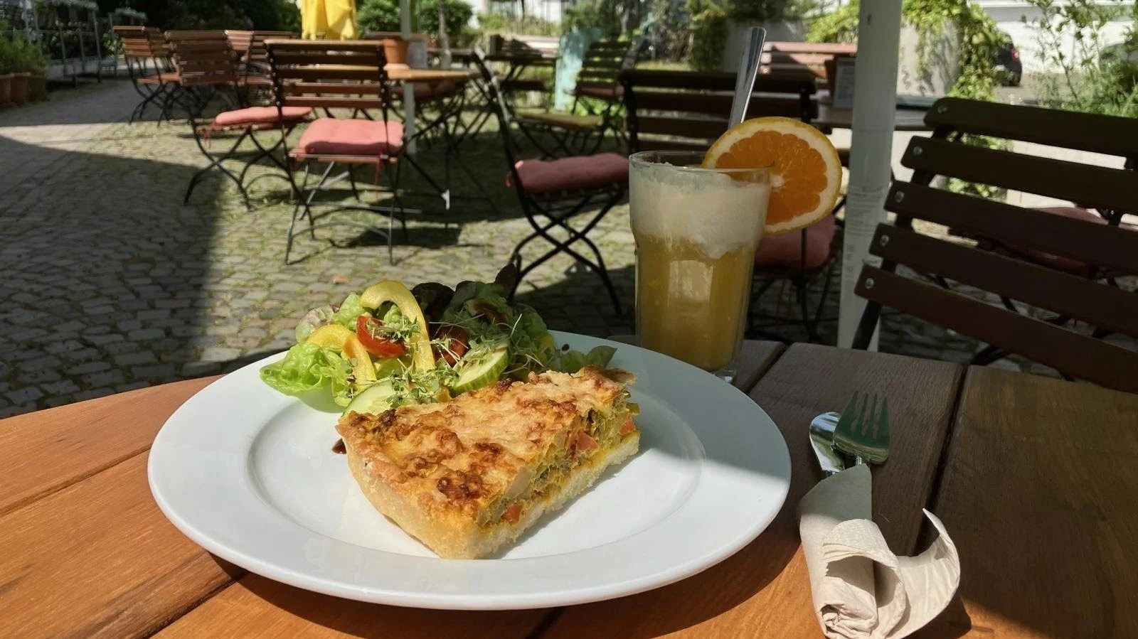 Ein Teller mit einem Stück Quiche und einem kleinen Salat, daneben ein Glas Zitronenlimonade mit einer Zitronenscheibe, auf einem Holztisch im Außenbereich eines Cafés bei schönem Wetter.