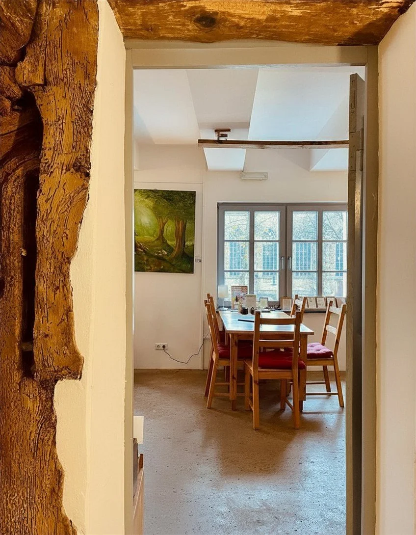 Ein Blick in einen hellen Speiseraum mit Holztischen und Stühlen, große Fenster mit Aussicht nach draußen, eine Wand mit einem grünen Baum-Balustrade-Balancier-​​Gemälde, Holzbalken an der Decke.