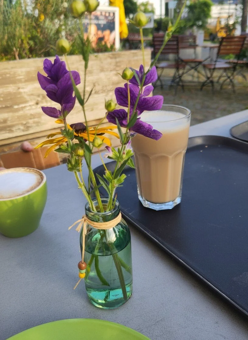 Blumen in einer kleinen Flasche auf einem Tisch mit Getränken, draußen bei Sonnenlicht.