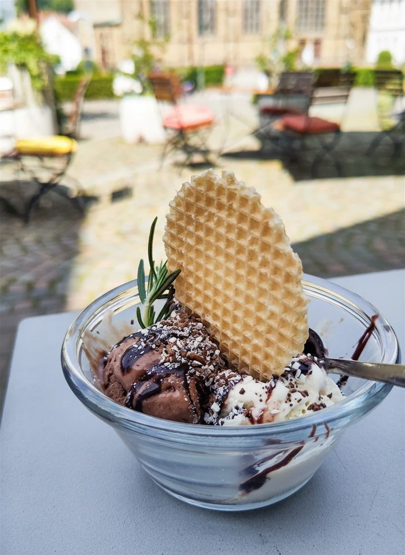 Eisschüssel mit Schokoladeneis, Vanilleeis, Waffel, Schokoladensauce, Rosmarin und Puderzucker, draußen auf einem Tisch.