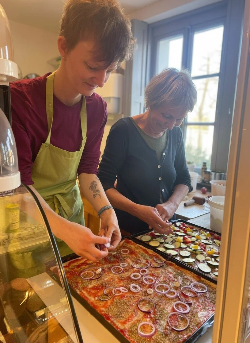Zwei Frauen beim Küchenbacken, eine mit grüner Schürze und die andere in dunkler Kleidung, bereiten eine Pizza mit Zwiebeln, Paprika und Zucchini im Ofen vor.